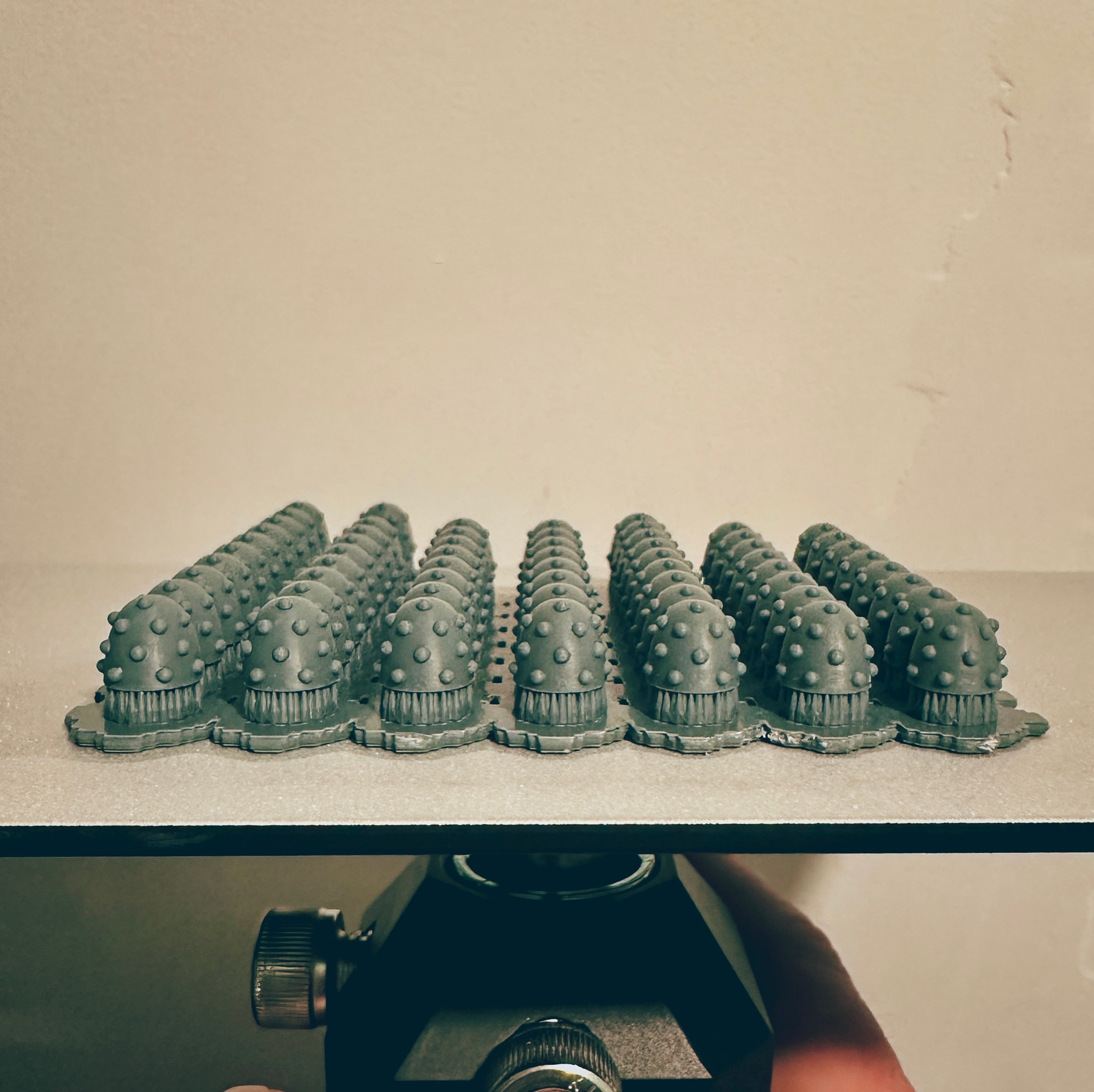 An upturned 3D printer build plate, showing a batch of 60 studded Space Marine shoulder pads, in the style of Mk 6 marines from the Horus Heresy: Age of Darkness boxset. They have been printed in a dark grey resin