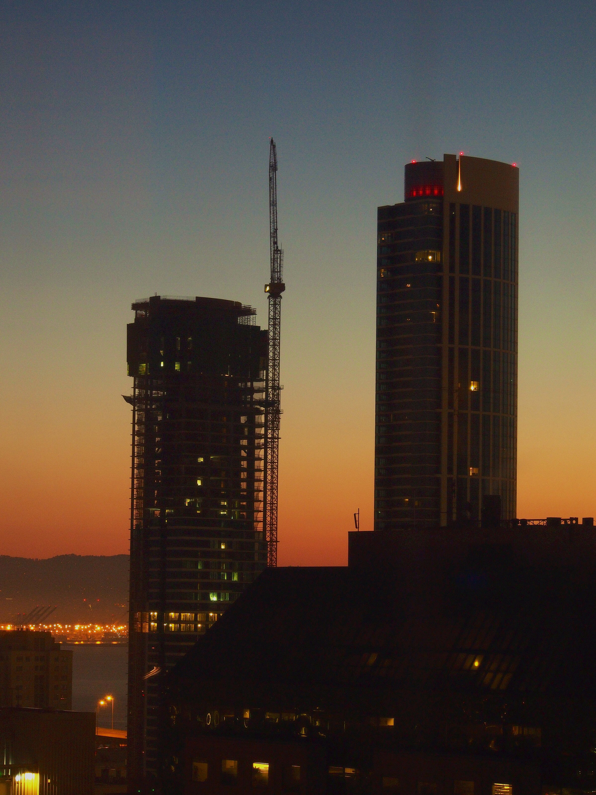 2 skyscrapers and a crane in San Francisco, framed by a sunrise sky which transitions from dark blue at the top to fiery orange near the bottom third. In the foreground is the silhouette of another building