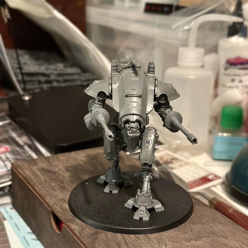 A detailed grey model of a bipedal robot stands on a circular black base. It is positioned centrally on a wooden surface, with its two gun arms raised, as if ready for action. It has a hunched over look due to the curving armoured carapace that rises over its head. The background features cluttered work surfaces with various tools and bottles, indicating a creative workspace.