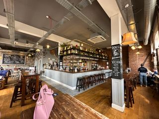 The interior of The Craftsman, taken from the corner of the room; with a bar area showcasing a variety of bottles, bar stools, and wooden dining tables with patrons. The ceiling has exposed ductwork, and there are blackboard menus with specials. A pink purse is on a table, and a “DOGS WELCOME” sign indicates a pet-friendly policy