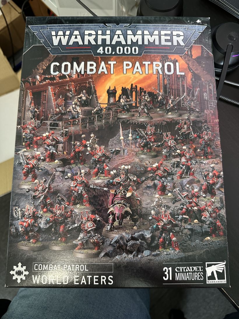 A boxed set titled 'Warhammer 40,000 Combat Patrol' showcases an array of miniature figures. The prominently featured red and brass miniatures depict various warriors, including one on a mounted steed, set against a dramatic and dystopian battlefield backdrop. The packaging indicates it includes 31 Citadel miniatures and is themed around the 'World Eaters.'