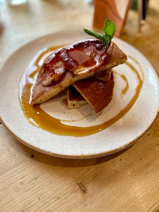 A pile of pancake halves and bacon, layered alternately, on a beige stoneware plate. Maple syrup has been poured on top of the food and then swirled around the plate to appear artful.