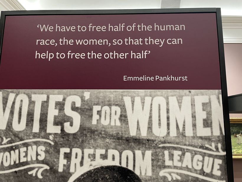 A sign displays a quote attributed to Emmeline Pankhurst, stating the necessity of freeing women to liberate society as a whole. The background features historical imagery related to women's rights, highlighted by the words “VOTES FOR WOMEN” and “WOMEN'S FREEDOM LEAGUE.” The display is part of an exhibition at Aberdeen Art Gallery related to the connection between women’s art and women’s liberation