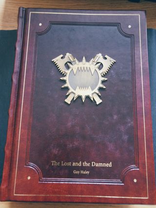 A red leather book cover, featuring embossing and a metal badge of the same design as was on the sticker. The title, The Lost and the Damned, and author name, Guy Haley, are picked out in gold lettering