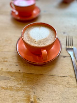 A oat-milk latte in an orange stoneware cup, on a matching saucer, placed on a rustic wooden table. The foamed milk has been poured in such a way as to resemble a heart.