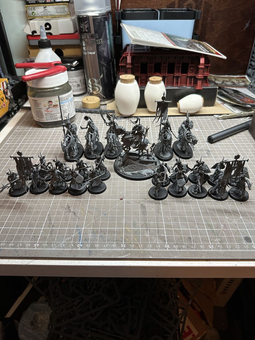 Skeletal figures are arranged on a cutting mat, showcasing various miniature models, predominantly in dark tone. A central figure stands larger, atop a broken tomb, indicating a commanding position, surrounded by smaller figures, some holding banners, and flanked by deathly knights on skeletal steeds. These miniatures are assembled, but unpainted, “Deathrattle” miniatures by Games Workshop, for their fantasy game Age of Sigmar. The backdrop includes various painting materials and tools, suggesting an active workspace for model painting or assembly.