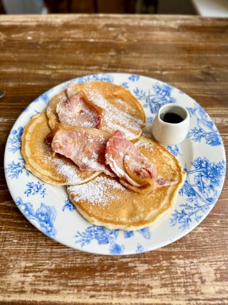 The image features a plate with three pancakes topped with bacon and powdered sugar, accompanied by a container of syrup. The plate has a blue floral design and rests on a rustic wooden table.