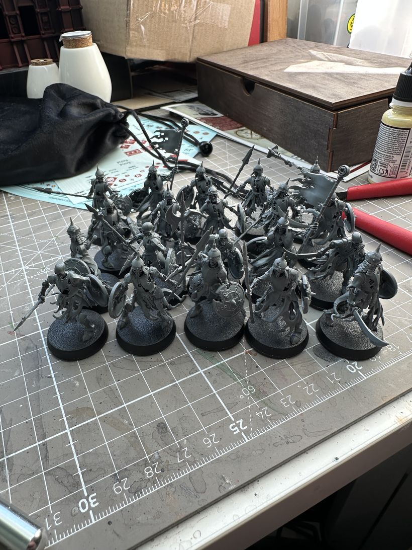 A collection of unpainted miniature figures, depicting warriors with various weapons and shields, is arranged in a cluster on a cutting mat. Surrounding them are tools and materials used for modelling, including a black drawstring bag, a wooden box, and bottles of paint. The setting suggests an area for hobby crafting.