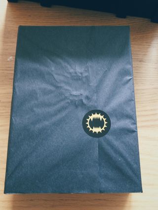 Black tissue paper wrapped around a book. The wrapping is secured with a sticker featuring a design evocative of a spiked collar containing a fanged maw.
