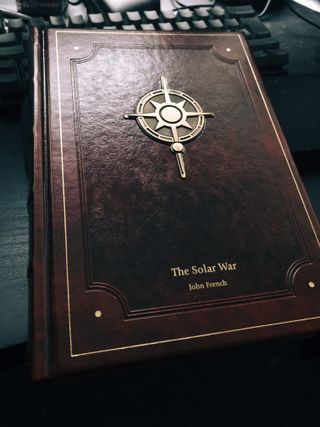 A red leather book cover, featuring embossing and a metal badge of the same compass/star design as was on the sticker. The title, Solar War, and author name, John French, are picked out in gold lettering