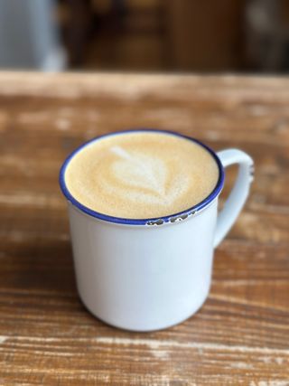 A oat-milk latte in an enamelled tin cup, placed on a rustic wooden table. The foamed milk has been poured in such a way as to resemble a heart. The blue enamelling around the rim has been chipped through frequent use, as has the handle.