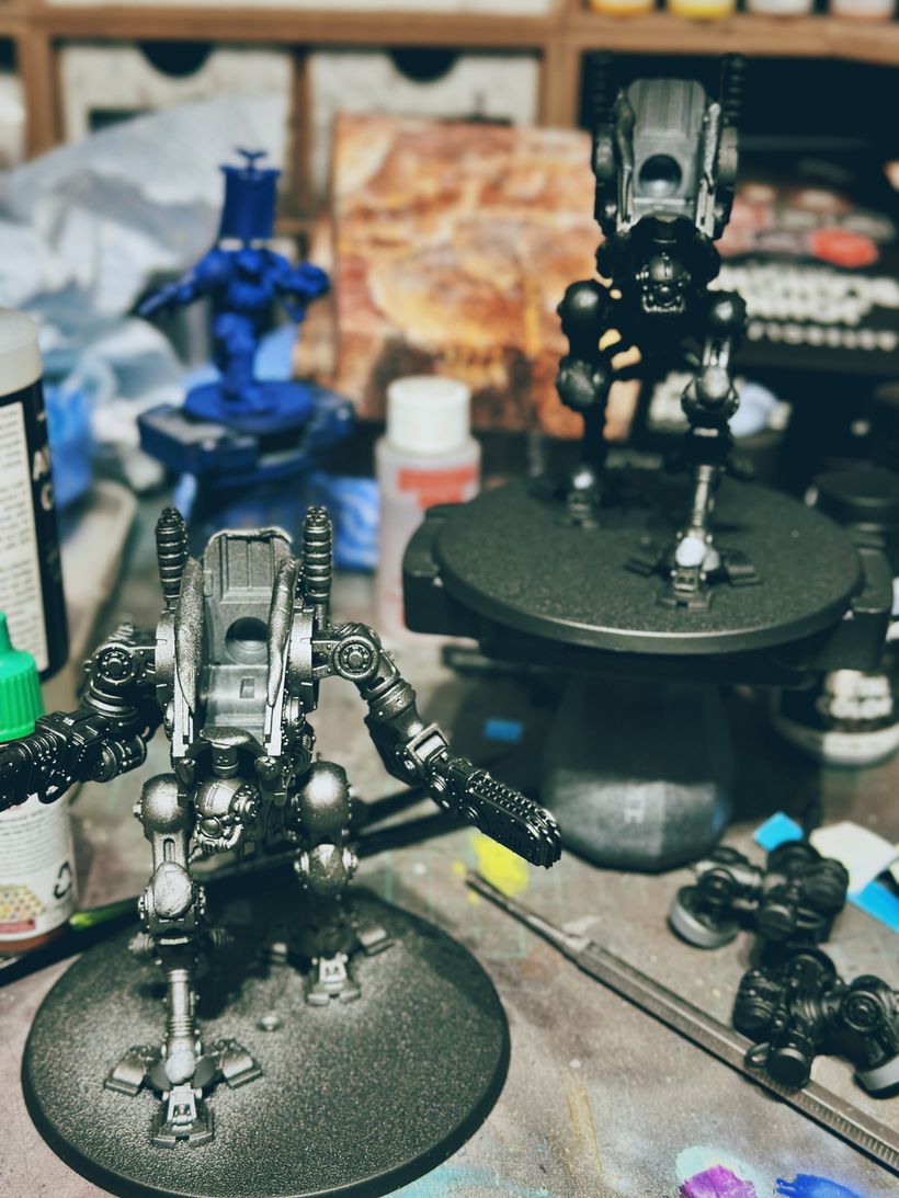 Two detailed robot figures stand on their bases, appearing partially painted in black primer and silver. One is positioned on the desk, while the other is on a painting handle. The backdrop features a blurred mix of tools, paint bottles, and an abstract artwork, suggesting a workshop environment dedicated to model painting.
