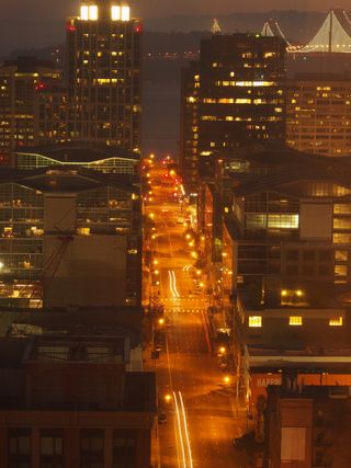 low-light, long exposure, photograph of Howard Street in San Francisco at sunrise, taken from a high vantage point. There are some light trails caused by passing cars. In the background is part of the bay, and the San Francisco-Oakland bridge.