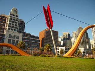 A large sculpture of a bow-and-arrow, sunk into the grassy ground, with the tip of arrow and some of the bow obscured by the ground. The fletchings of the arrow are bright red, and its shaft is painted white. The bow is a golden yellow colour. In the background are palm trees, some buildings of the San Francisco skyline, and a bright blue sky