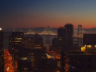 The San Francisco-Oakland bridge at sunrise. The bridge is illuminated with lights to show off the support cables. In the foreground are the silhouettes of various buildings and some streets.