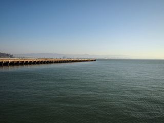 A pier jutting out into the water of the San Francisco bay, from the left side of the frame. In the hazy distance, hills can be seen