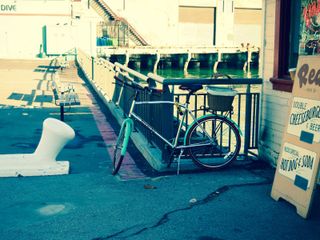 A vintage style bicycle, padlocked to a railing next to Red's Java House in San Francisco. In the background is a pier building, and to the right is a partial view of a sign advertising Double Cheeseburger & Beer, and Red's Special Hot-Dog and Soda