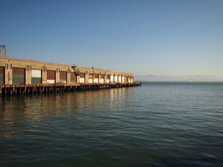 A pier jutting out into the water of the San Francisco bay, from the left side of the frame. In the hazy distance, hills can be seen. The pier has a building with several warehouse doors on it.