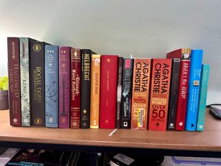 A row of 17 books, mainly hardback fiction, on a wooden sideboard top